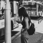 Black and white photo of a woman at a city bus stop, using her phone and wearing casual attire.