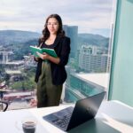 Businesswoman in office holding notebook by window overlooking cityscape.